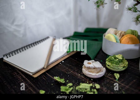 Zerkleinert, Pastelle, orange und grüne Makronen auf Holz Hintergrund mit ganzen in weißen Teller mit Krug mit Blumen auf der Rückseite Stockfoto