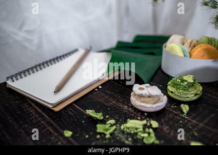 Zerkleinert, Pastelle, orange und grüne Makronen auf Holz Hintergrund mit ganzen in weißen Teller mit Krug mit Blumen auf der Rückseite Stockfoto