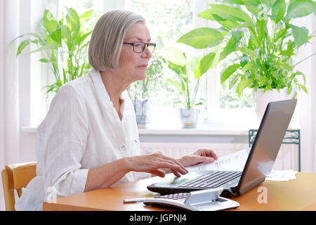 Entspannte senior Frau zu Hause mit einem Taschenrechner und viele Belege vor ihrem Laptop machen ihre jährliche Steuererklärung Stockfoto