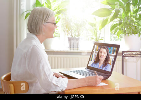 Ältere Frau zu Hause vor ihrem Laptop Notizen während gerade eine Online-Englisch-Lektion von einem weiblichen Lehrer, e-Learning-Konzepts Stockfoto