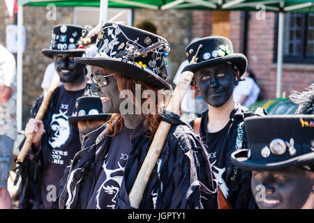 Traditionelle englische Volkstänzer, Hunters Moon Morris Dancers, Nahaufnahme der lächelnde Frau mit geschwärzt Gesicht, schwarzer Hut und t-shirt, Teenager hinter ihr auf Viewer, Auge - Kontakt. Stockfoto