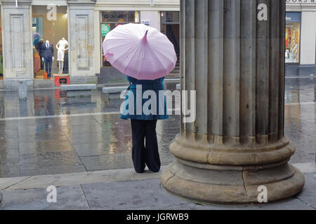 junges Mädchen Teenager Regen in Glasgow mit rosa Regenschirm Sonnenschirm auf die Straße nass Regen Tag neben Spalte brollie Stockfoto