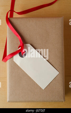 Overhead Schuss eine verpackte Packpapier-Geschenk-Paket auf Holzfurnier Tisch. Gekrönt mit einem Paket Tag und rotes Band, zeigt die leere Beschriftung nach oben, pr Stockfoto