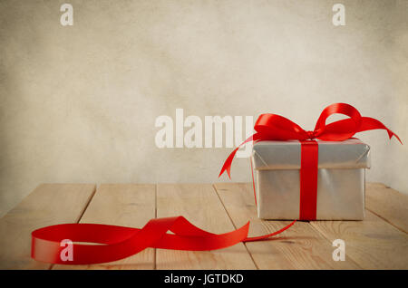 Eine Weihnachts-Geschenk-Box mit geschlossenem Deckel in Silber Papier eingewickelt und an einen Bogen mit einem roten Satinband gebunden.  Platziert auf einem verwitterten alten Holztisch mit Stockfoto