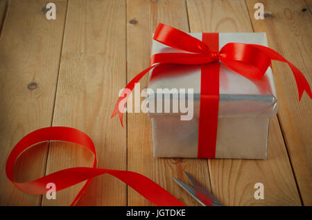 Geschenk-Verpackung-Szene. Eine Weihnachts-Geschenk-Box in Silber Papier gewickelt und gebunden mit roten Band zu einer Schleife auf eine alte Holzbohle Tabelle mit Schere und Re Stockfoto