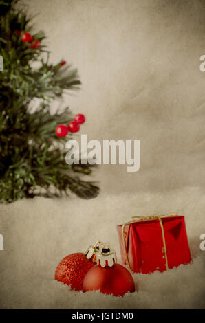 Rote Weihnachtskugeln eingebettet in Dekoschnee mit Weihnachtsbaum im Hintergrund beladen mit Beeren.  Grunge Vintage aussehen. Stockfoto