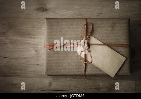 Overhead Schuss eine Geschenkbox mit einem Grunge Paket Tag für ein Label, verpackt in braunem Papier mit einem Bast Band gebunden an einen Bogen auf geplankte Holztisch. Stockfoto