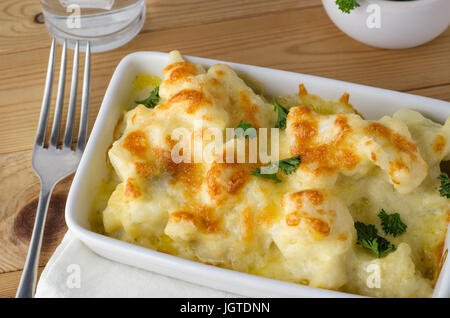 Gastro-Szene. Eine Schüssel mit goldenen gebackenen Blumenkohl Käse, verstreut mit frischer Petersilie Blätter.  Silber Gabelung links auf Kiefer Plank Holztisch.  Nur vis Stockfoto