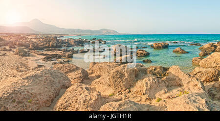 Panorama-Bild der Sonne hinter Bergen auf Falasarna Strand und Beleuchtung Felsen und Mittelmeer mit warmem Licht, Kreta, Griechenland Stockfoto