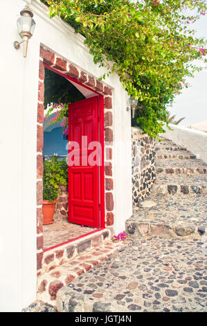 Rote offene Tür eines Hauses mit blühenden Pflanze drüber an warmen Frühlingstag mit Boulder Treppen in Oia, Santorini oder Thira, Griechenland Stockfoto