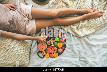 Sommer gesunde vegane sauber Essen Frühstück im Bettenkonzept. Junges Mädchen Kleidung Pastell farbigen nach Hause nehmen Obst aus Tablett voller Frische se Stockfoto