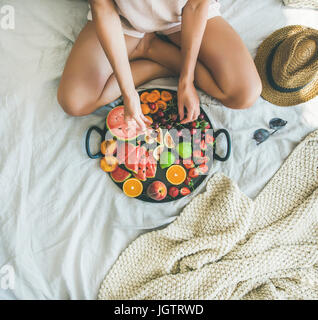 Sommer gesunde vegane sauber Essen Frühstück im Bettenkonzept. Junge Mädchen tragen Pastell gefärbt nach Hause Kleider nehmen Kirschen aus Fach voller Frische Stockfoto