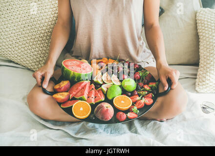 Sommer gesunde vegane sauber Essen Frühstück im Bettenkonzept. Junge Mädchen tragen Pastell gefärbt nach Hause Kleider sitzen und halten Tablett voller Frische Stockfoto