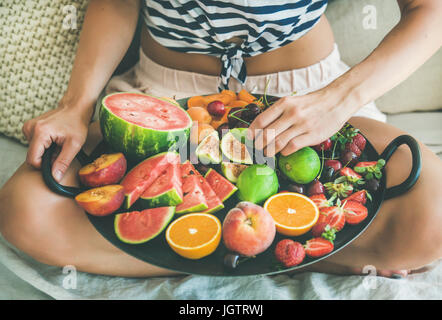 Sommer gesunde vegane sauber Essen Frühstück im Bettenkonzept. Junge Mädchen tragen gestreifte Heimtrikot sitzen und nehmen Obst aus Tablett voller Frische Stockfoto