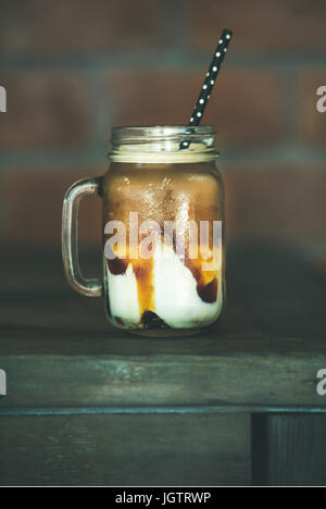 Iced Caramel Macciato Milchkaffee im Glas mit Strohhalm auf dunklen Holztisch, selektiven Fokus. Kalte süße erfrischende Sommer Getränk Konzept Stockfoto