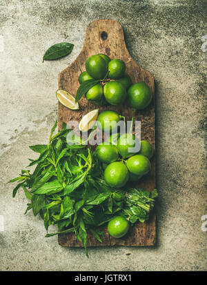 Flatlay von frisch gepflückten Bio Limetten und Minze Blätter für Herstellung cocktail oder Limonade auf rustikalem Holzbrett über grauen Beton Stein Hintergrund zu Stockfoto