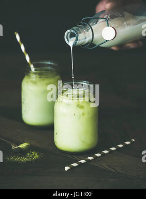 Kalten Erfrischungsgetränk Geeiste Kokosnuss Matcha Latte, hand Gießen Milch aus der Flasche. Tiefenschärfe, dunklen Hintergrund. Sauber, Essen, gesunde, vegetarische, v Stockfoto