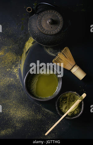 Grüner Tee Matcha-Pulver und Heißgetränk im schwarzen Schalen stehen mit Eisen-Teekanne, traditionelle Werkzeuge Bambuslöffel und Schneebesen über dunklem Metall. T Stockfoto