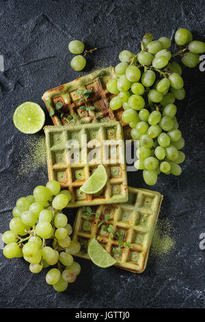 Hausgemachte quadratische grünen Matcha belgische Waffeln mit frischen reifen Trauben, Minze und Limette über schwarze Steinstruktur Hintergrund. Draufsicht mit Raum Stockfoto