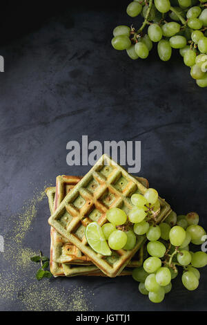 Hausgemachte quadratische grünen Matcha belgische Waffeln mit frischen reifen Trauben, Minze und Limette über schwarze Textur Metall Hintergrund. Draufsicht mit Raum Stockfoto