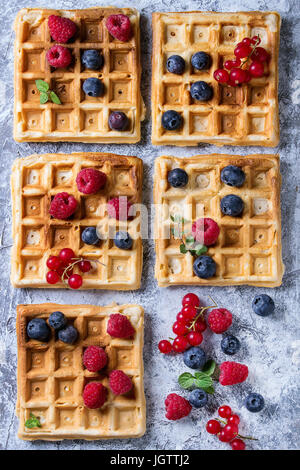 Hausgemachte quadratische Waffeln mit frischen reifen Beeren Heidelbeeren, Himbeeren, Johannisbeeren über graue Textur Hintergrund. Draufsicht mit Raum Stockfoto