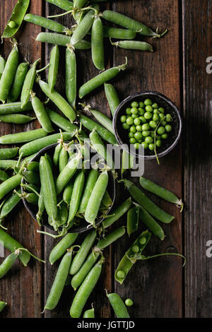 Junge organischen grünen Erbsenschoten und Erbsen in Schalen über alten dunklen hölzernen Planken Hintergrund. Ansicht von oben mit dem Raum. Ernte, gesund essen. Stockfoto