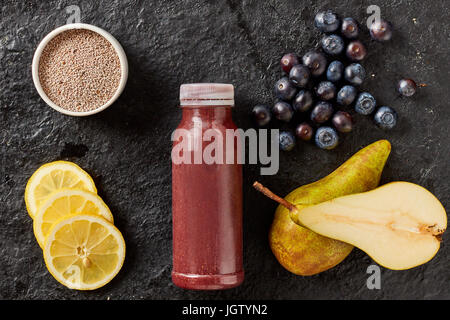 Frische Frucht-Smoothie mit Heidelbeeren, Zitrone und Birne mit Chiasamen vermischt und serviert in einer Flasche, umgeben von den Zutaten auf einem strukturierten schwarzen Stockfoto