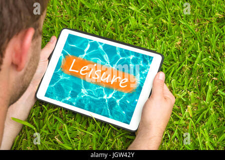 Junger Mann liegend auf dem Rasen im Garten mit Blick auf Wasser Hintergrund mit Wort "Freizeit" darauf geschrieben Tablet-Gerät. Stockfoto