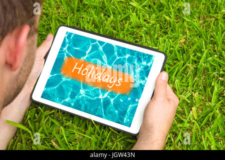 Junger Mann liegend auf dem Rasen im Garten mit Tablet-Gerät betrachten Wasser Hintergrund mit Wort "Urlaub" darauf geschrieben. Stockfoto