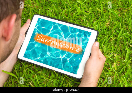 Junger Mann liegend auf dem Rasen im Garten mit Blick auf Wasser Hintergrund mit Wort "Summertime" darauf geschrieben Tablet-Gerät. Stockfoto