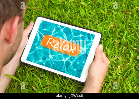 Junger Mann liegend auf dem Rasen im Garten mit Blick auf Wasser Hintergrund mit Wort "Relax" darauf geschrieben Tablet-Gerät. Stockfoto