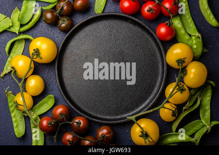 Essen-Hintergrund mit Textfreiraum. Rote und gelbe Tomaten und grüne Erbse um Epty schwarz Guss Eisenplatte. Ansicht von oben Stockfoto