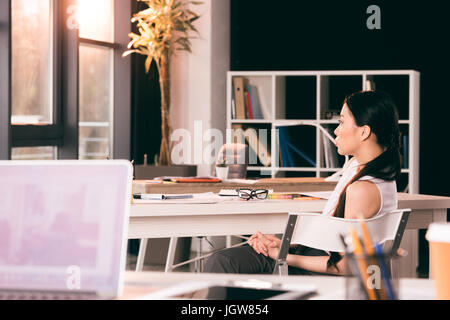 junge asiatische Geschäftsfrau sitzt am Tisch im Büro und auf der Suche zur Seite Stockfoto