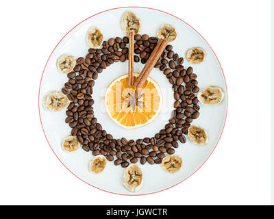 Körner von geröstetem Kaffee, Tassen getrocknete Orange und Banane, Zimtstange, Anis Sterne liegen auf einem Porzellanteller in Form einer Uhr in eine Schüssel-isolat Stockfoto