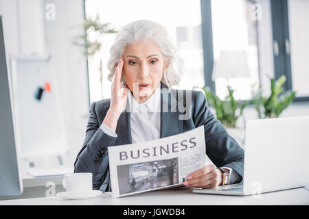 Schockiert senior Geschäftsfrau in Abendgarderobe Zeitungslektüre am Arbeitsplatz Stockfoto