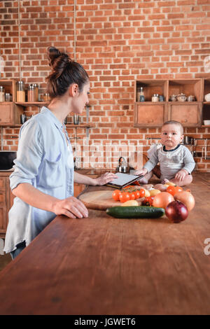 Porträt der jungen Mutter mit ihrem kleinen Sohn kochen Abendessen zusammen und mit digital-Tablette in der Küche Stockfoto