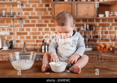 Entzückende kleine Junge spielt mit Mehl und sitzen am Küchentisch. Inländische Lebenskonzept Stockfoto