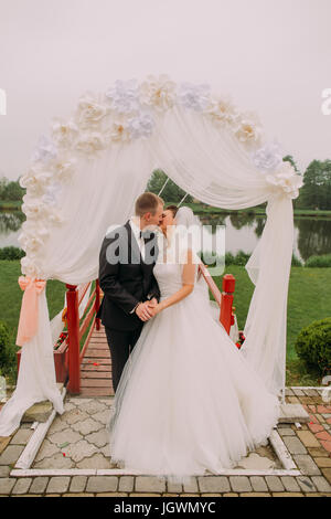 Das küssende Brautpaar unter dem Bogen Hochzeit. Das Full-length Foto. Stockfoto