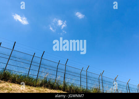 Ein Rahmen mit einer doppelten Draht-Zaun mit Stacheldraht auf einer Bank unter einer sengenden Sonne und blauen Sommerhimmel gemacht. Stockfoto