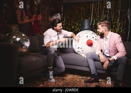 Zwei Männer sitzen auf Sofa bei Party, riesiger Schneemann Kopf dazwischen, lachen Stockfoto