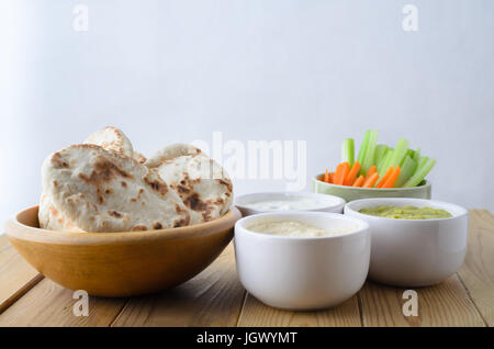 Eine Auswahl an Dips auf einem Holzbrett-Tisch, mit Schüsseln mit Fladenbrot und Crudites.  Enthält; Hummus; Guacamole; Käse und Schnittlauch; Sellerie und Karotte. Stockfoto