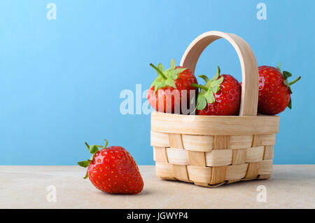 Frische rote Erdbeeren. gestapelten hoch und füllt einen kleinen geflochtenen Korb auf einer Holzfläche vor hellblauem Hintergrund. Stockfoto