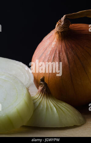 Nahaufnahme einer ganze, ungeschälte Zwiebel neben geschälten Scheiben auf ein Schneidbrett aus Holz mit schwarzem Hintergrund. Stockfoto