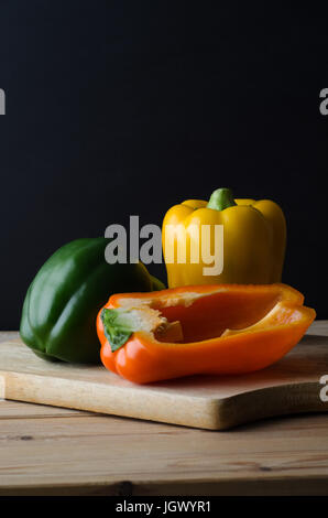 Ein Stillleben Anordnung der drei Paprika (grün, gelb und Orange) auf ein Schneidbrett aus Holz mit geplankte Küchentisch unten und schwarzen backg Stockfoto
