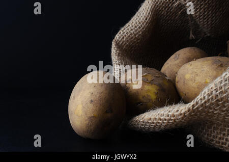 Roh, ungewaschen, ungeschälte Kartoffeln aus hessischen Sack auf schwarze Schieferoberfläche.  Stimmungsvolle Beleuchtung, schwarzer Hintergrund. Stockfoto