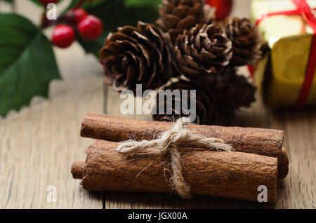 Zimtstangen, gebunden in einem Bundle mit Schnur an einem Eichentisch mit Weihnachten Holly, Tannenzapfen und ein Gold verpackt Geschenkbox im Hintergrund. Stockfoto