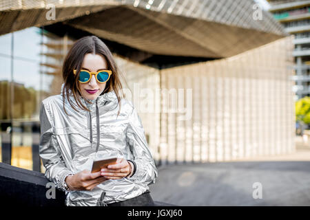 Lifestyle-Porträt einer stilvollen Frau in Silber Jacke mit Telefon im Freien auf dem modernen architektonischen Hintergrund Stockfoto