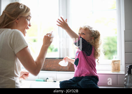 Mutter und Tochter in der Küche arbeiten Tochter sitzen auf Oberflächen mit Abwasch Bubbles spielen Stockfoto