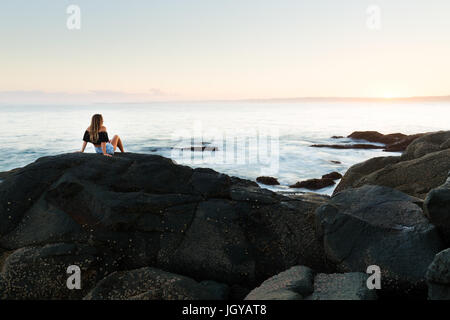 Ein gesundes junges Mädchen sitzt auf einem Stein und beobachtet den Sonnenuntergang über einem herrlichen Seelandschaft in Australien.