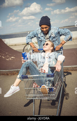 Jungen tragen fröhliches Mädchen mit kann im Warenkorb in der Nähe der Rampe am Skateboard-park Stockfoto